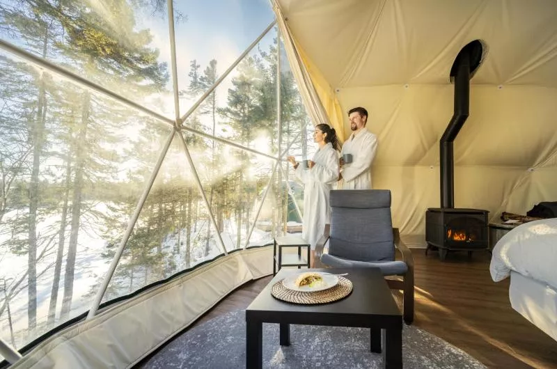 Two people wearing robes and holding coffee mugs stand in the window of a dome at White Point Beach Resort. There is a small fire on in the stove behind them.