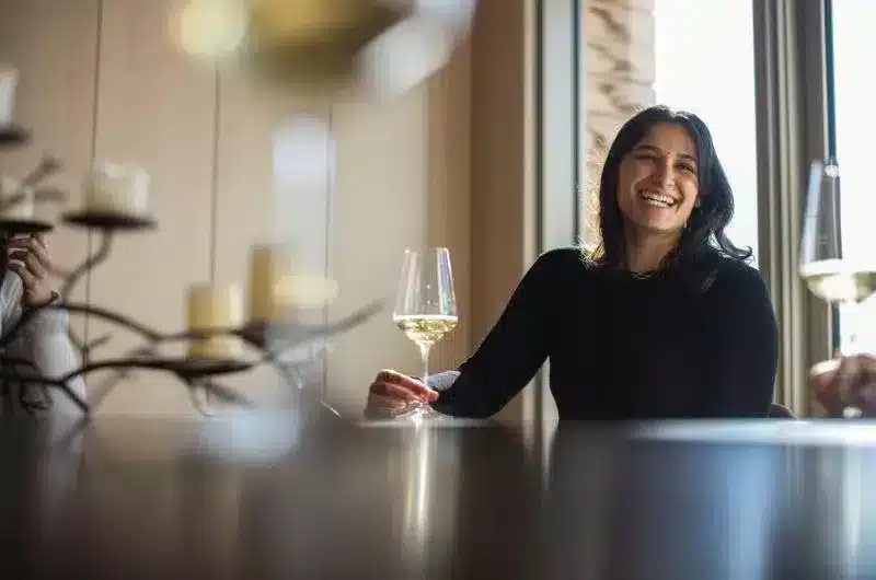 A person smiles while sitting at a table and holding a glass of white wine.