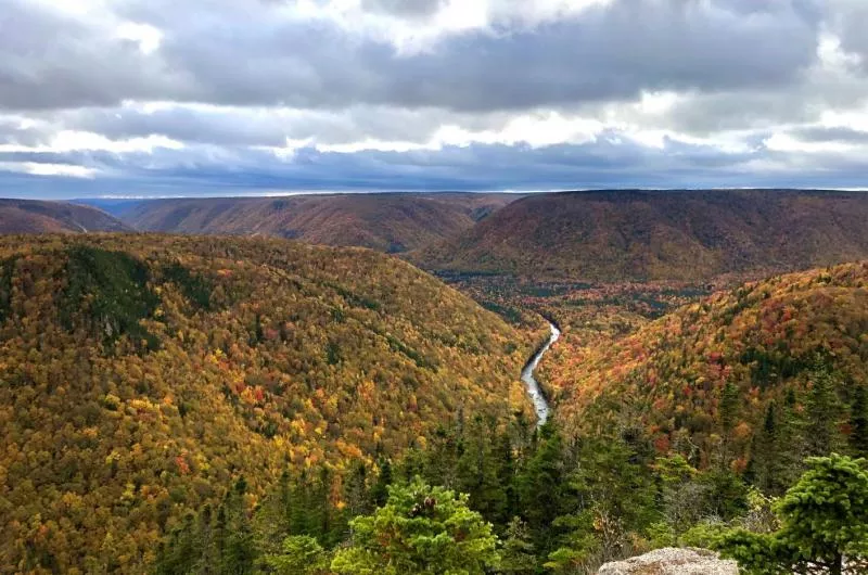 Several hills and valleys are dotted with trees that are changing into fall colours. At the bottom of the hills there is a river that winds through the photo. It is a sunny day but there are lots of fluffy clouds in the sky.