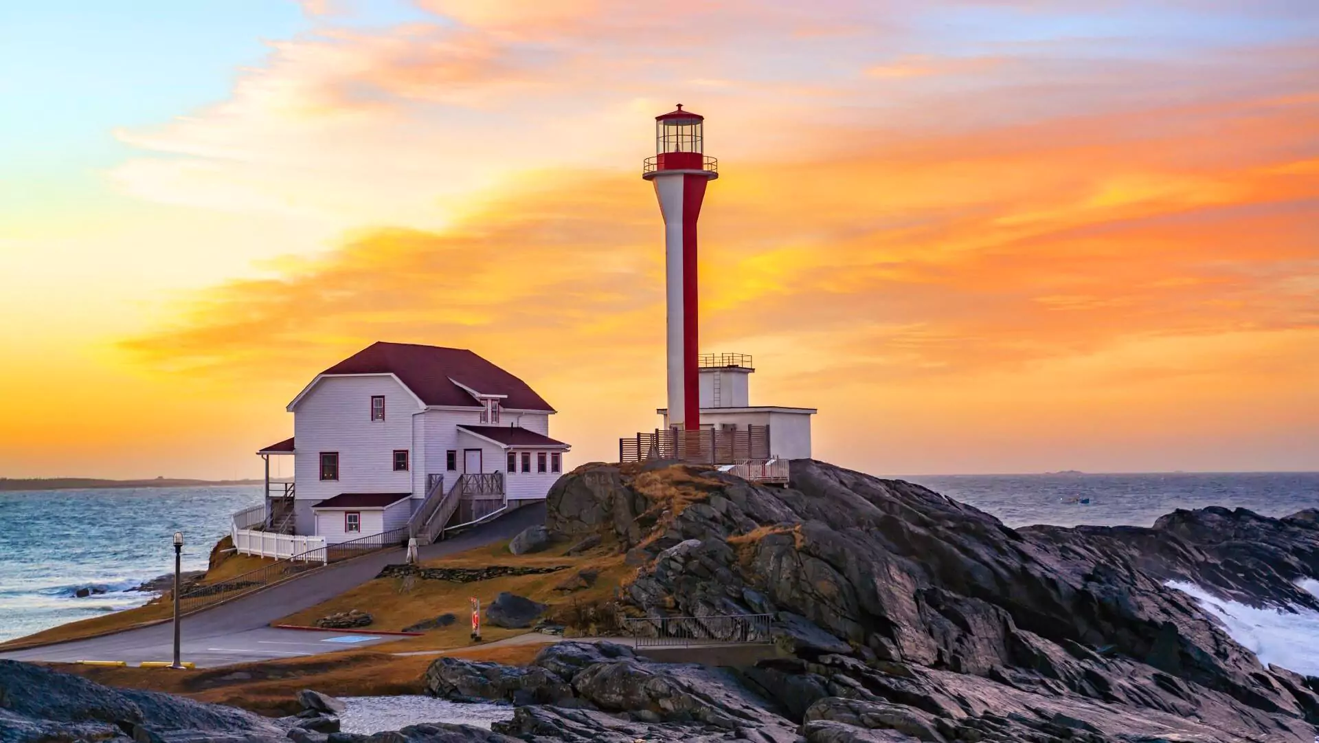 A red and white lighthouse sits atop rocks along the coast at sunset