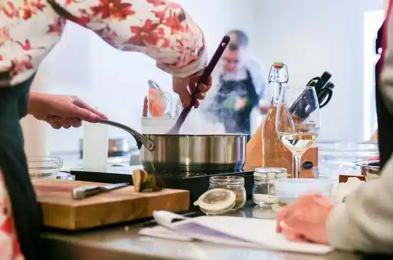 A person, only visible by the arms, stirs a pot with steam rising from it. They are clearly standing at a counter cooking with some ingredients visible on the counter.