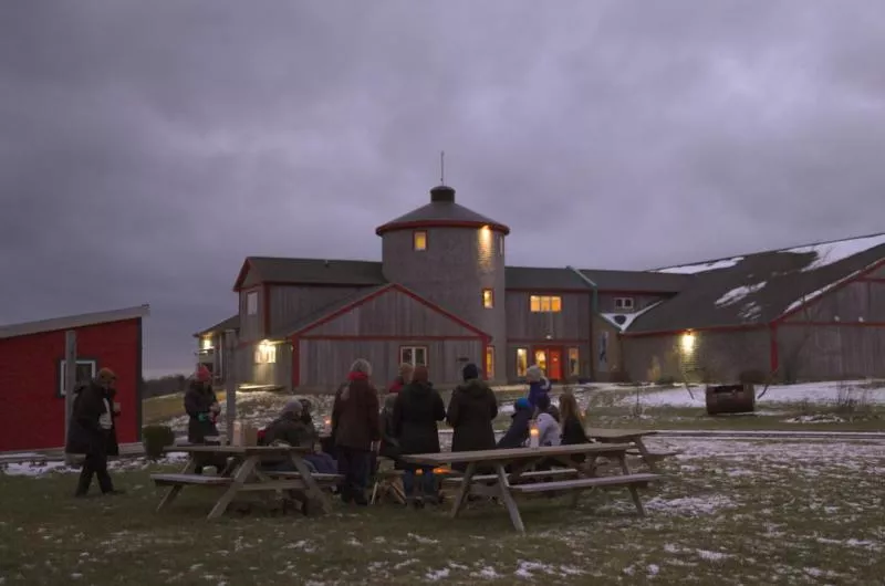 A group of people sit at picnic tables with their backs to the camera. It is winter and they are standing in front of a very large barn.