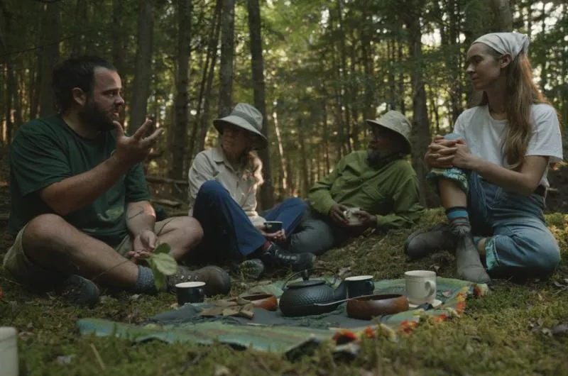 Four people sit in the forest, during summer. They are talking animatedly and there is a picnic blanket with food on the ground.