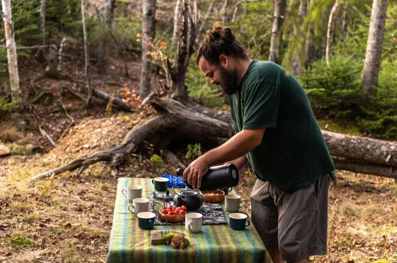 One person is pouring hot water from a thermos into a teapot at a table set for a group of people to eat. The setting is in the woods during fall.