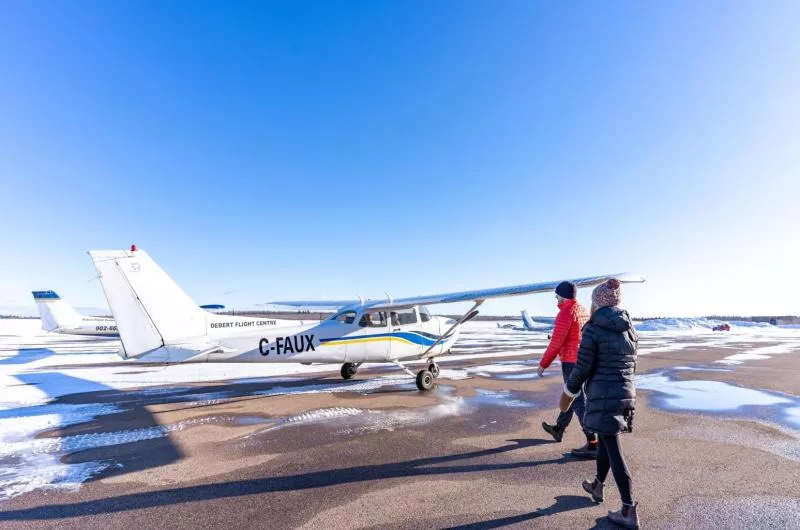 Two people walk toward a small plane on a landing strip. There is snow on the ground, but it is a blue sky day with no clouds.