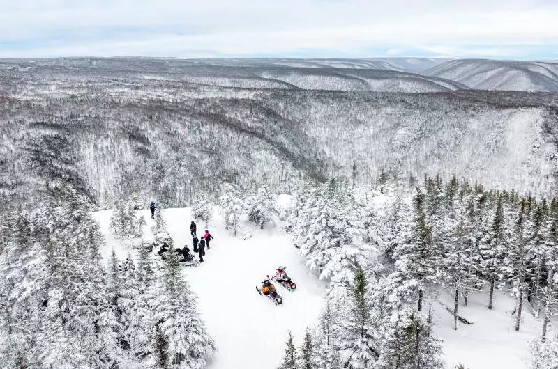A group of snowmobilers are stopped by the edge of a cliff. Everything is covered in snow and it is very picturesque.