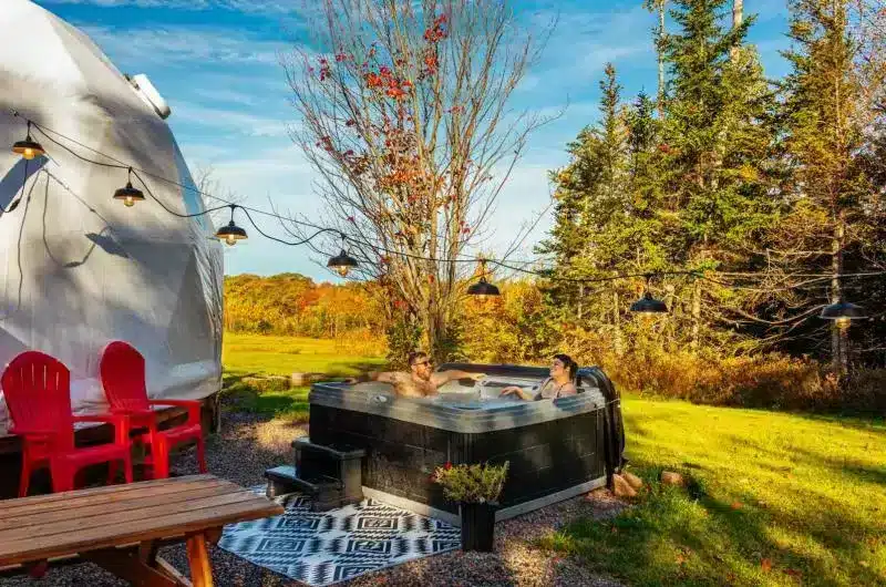 Two people sit in a hot tub outside a dome. It is fall and the trees are beginning to change colour.