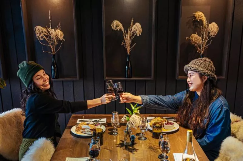 Two people cheers glasses of wine over a large wooden table. They both have meals in front of them.