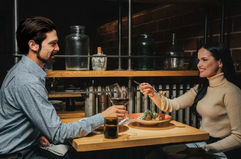 Two people are dining together at a restaurant. One is lifting a glass to drink wine while the other is eating pasta.