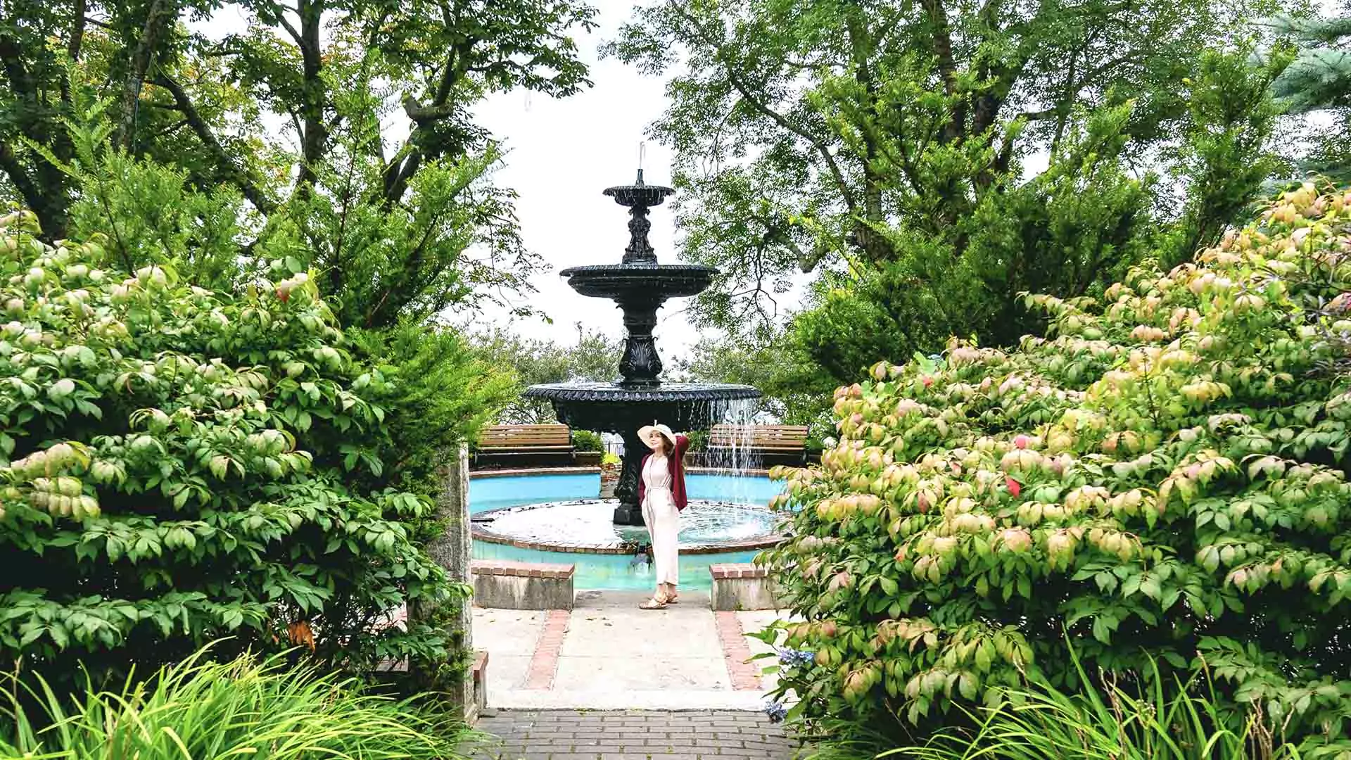 A person stands in front of the large fountain in the center of Frost Park in downtown Yarmouth, Nova Scotia, surrounded by lush greenery in summer