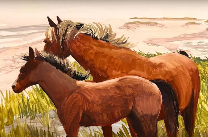 Two brown horses graze on green grass. In the background is lots of sand.