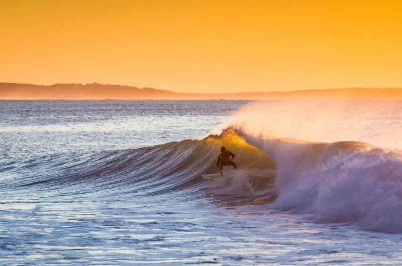 A person surfs a large wave during sunset. The sky is golden.