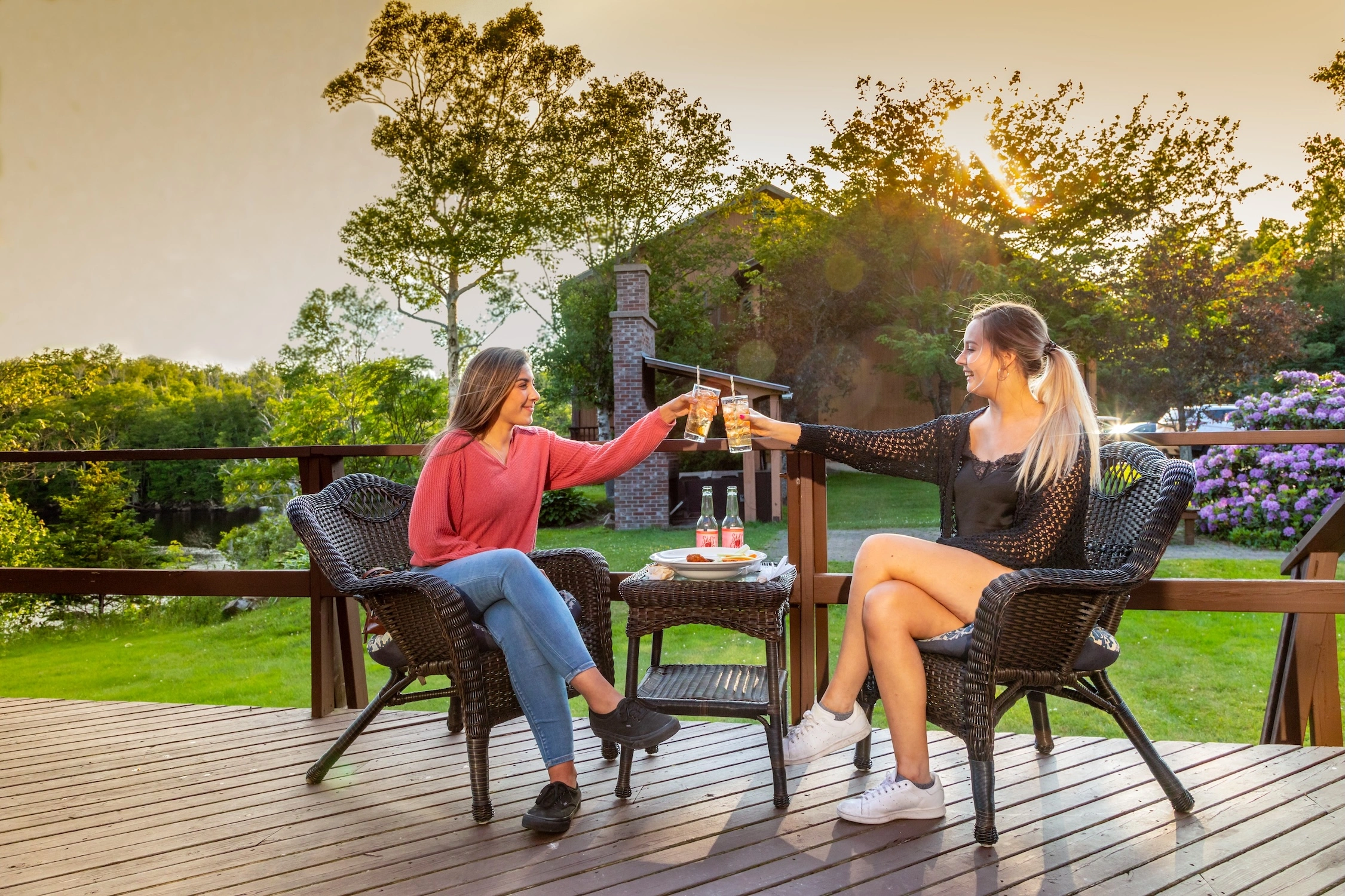Two women sitting on a deck with patio furniture having drinks and food