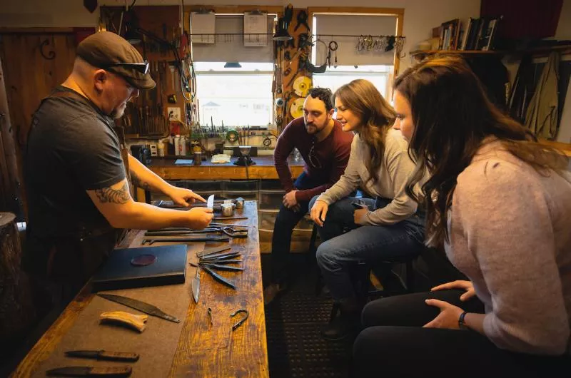 A group of people take part in a knifemaking demonstration. They sit in front of the instructor at a table, inside a workshop