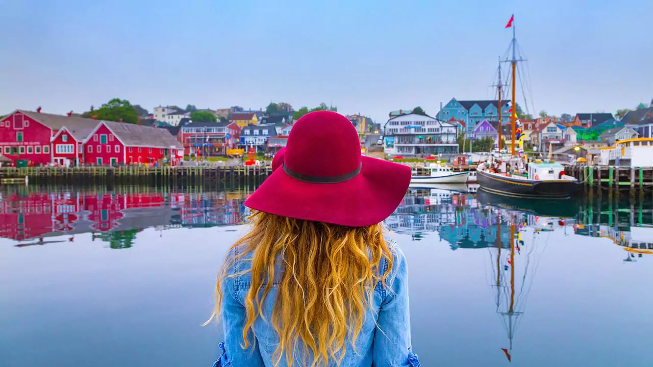 A lady in a blue shirt and red hat with blond flowy hair, stands in front of the Lunenburg waterfront which is full of colourful buildings and boats in the harbour