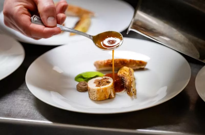 A hand spoons a sauce onto a piece of meat and vegetable on a white plate.