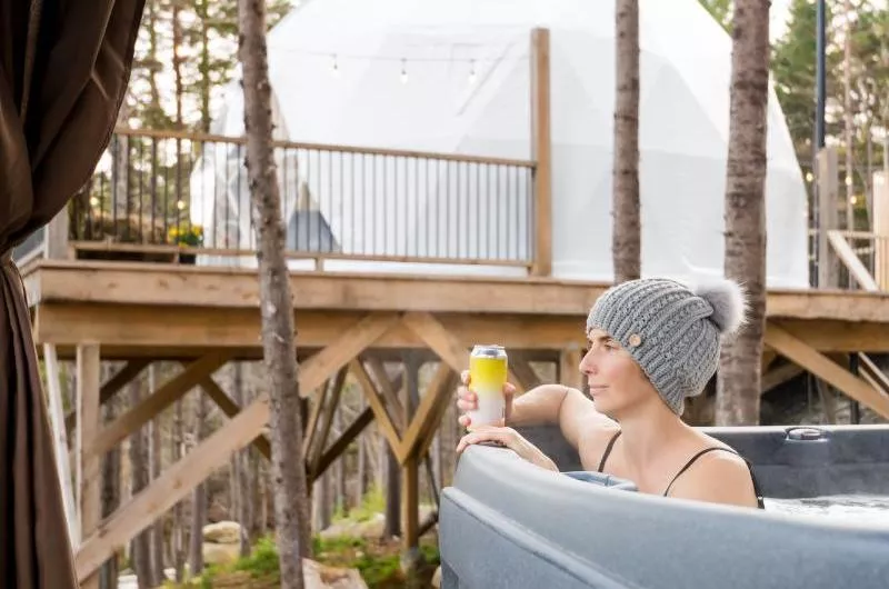 A person sits in a hot tub with a canned beverage. They are wearing a winter hat, indicating it is old out. In the background there is a white dome.