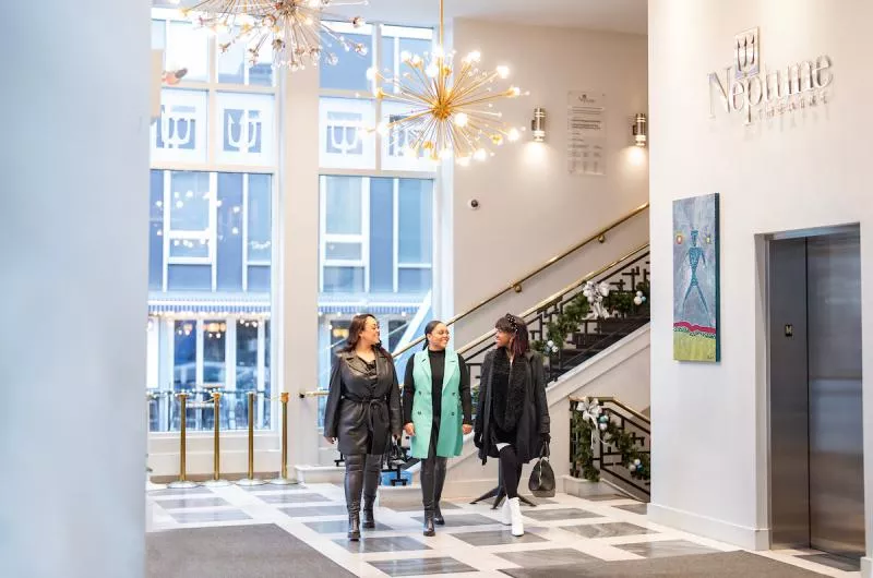 Three people inside the lobby area of Neptune Theatre in Downtown Halifax. It is brightly lit with lots of chandeliers.