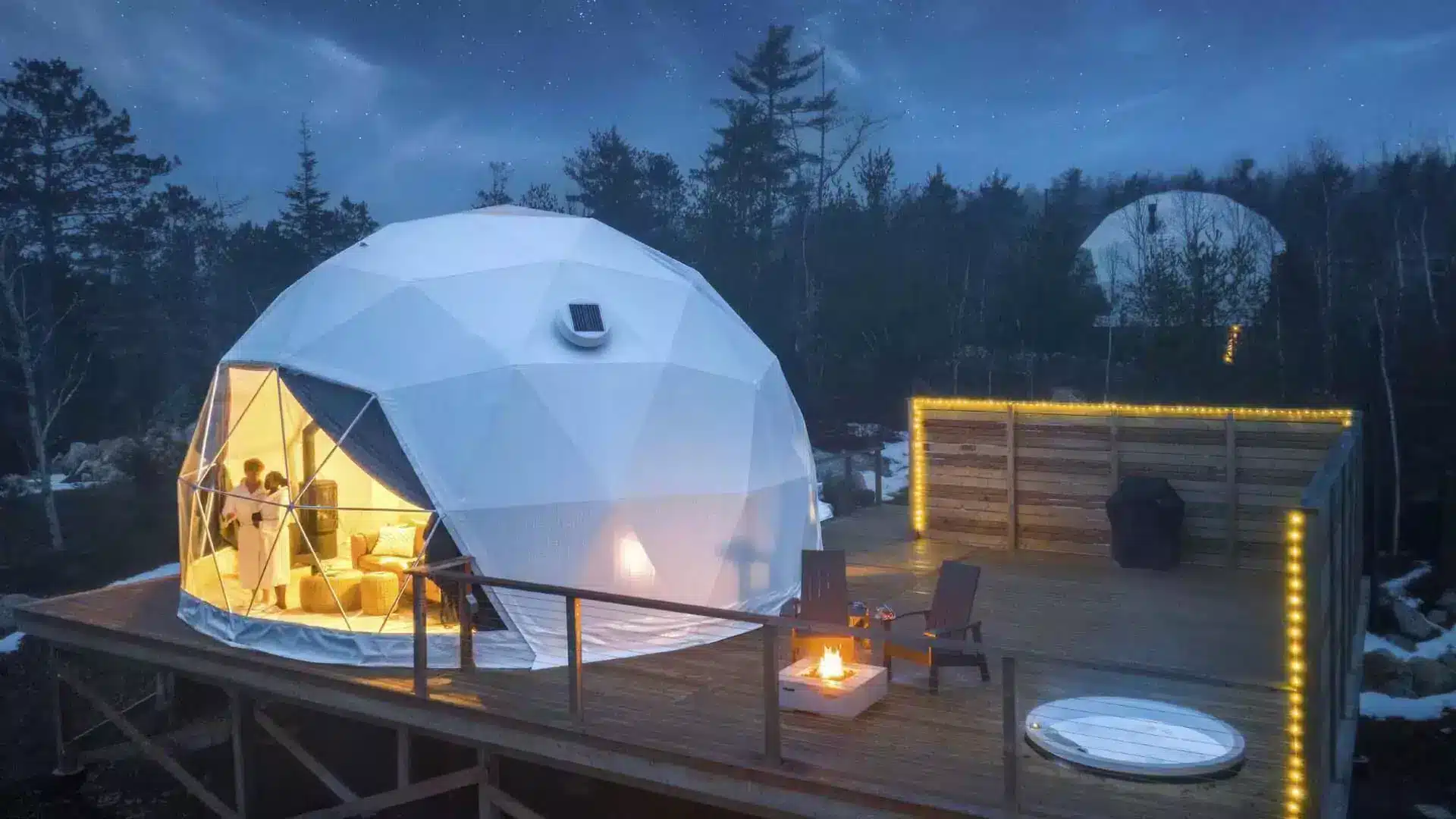 A geodesic dome is lit from the inside at dusk with a hot tub next to it on the deck