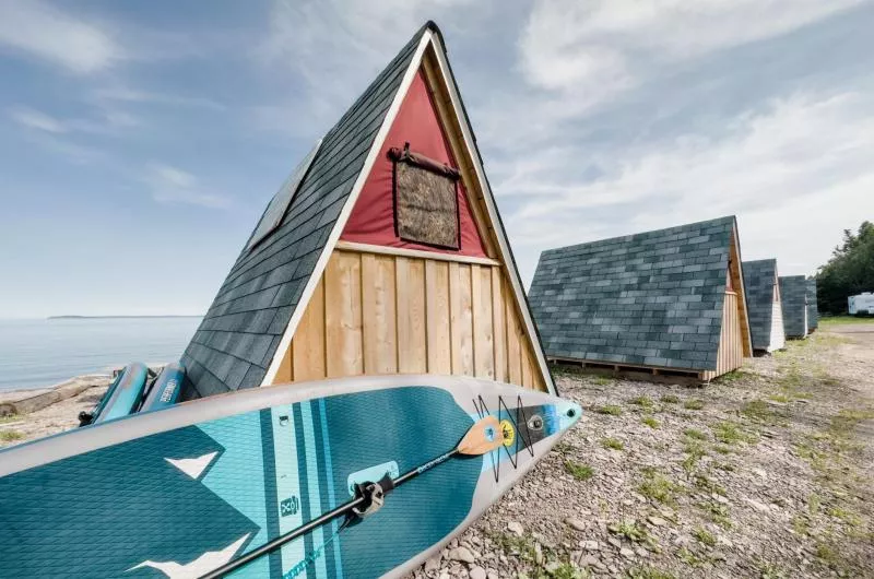 A wooden a frame (tent) sits on the sand on the edge of the Bay of Fundy's waters with a turquoise paddleboard perched against it