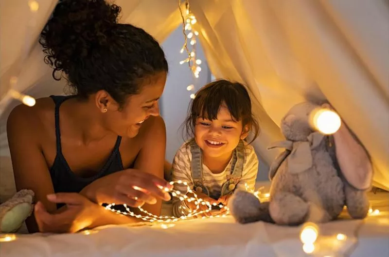 A mother and child lay on their stomachs in what appears to be a tent with lots of twinkle lights.