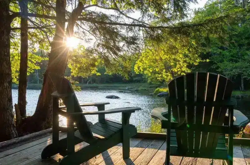Two Adirondacks appear to sit on a deck near a lake. It appears to be summer and the trees are very lush.