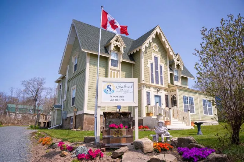 Exterior of the Seabank House Bed and Breakfast. There is a sign in front with the name. There is a Canadian flag. The siding on the older style home is a pale green/taupe. There are flowers out front.