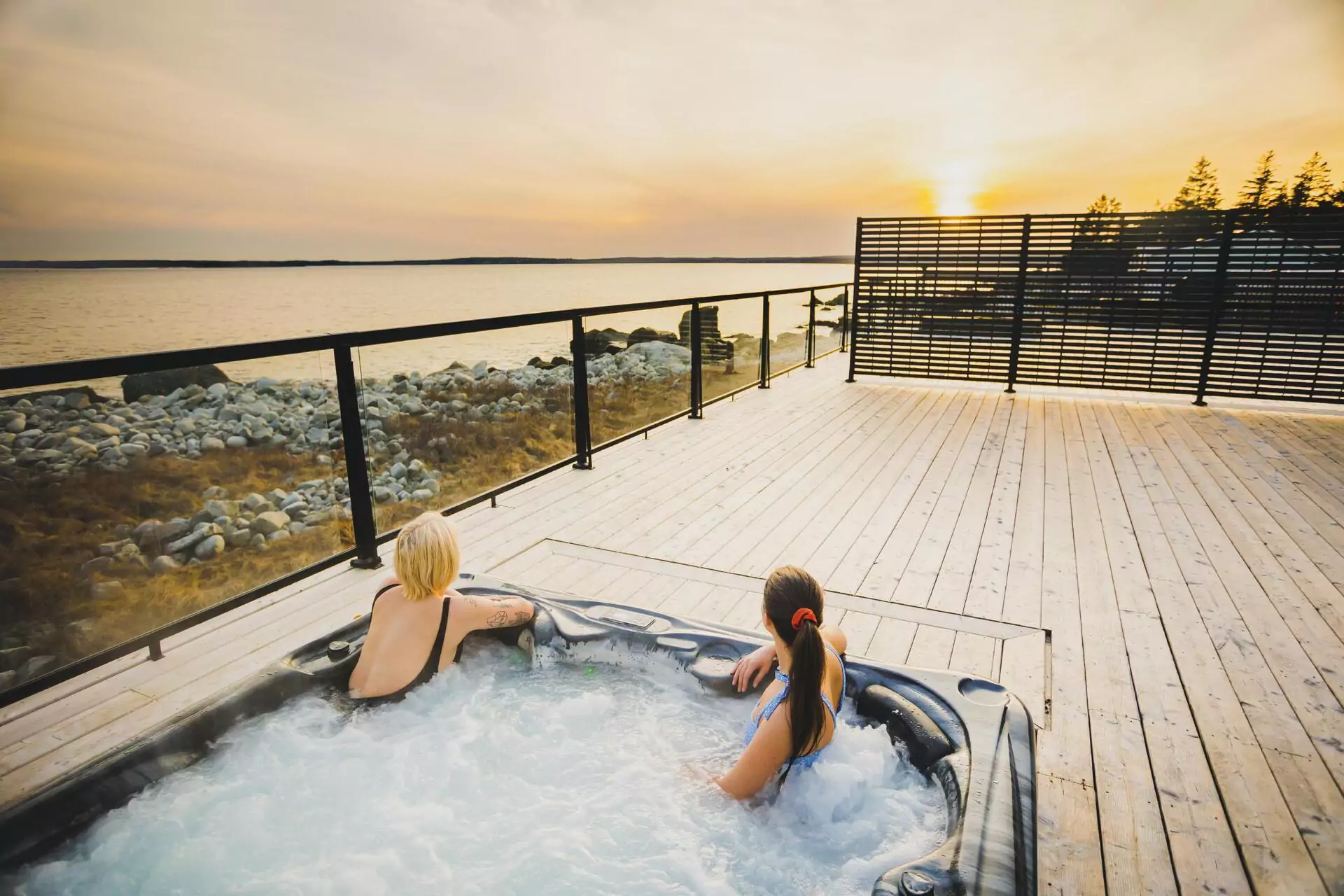 Two women relax in a hot tub on a large deck overlooking a beach as the sun sets.
