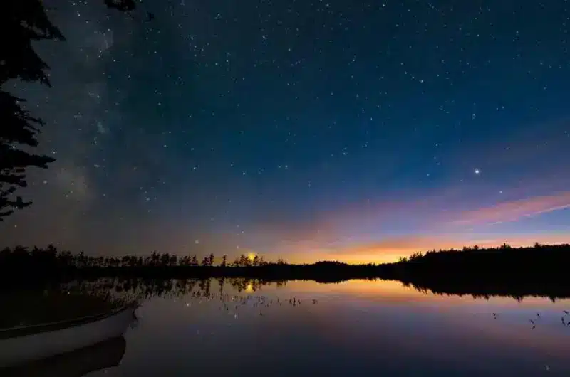 A sunset with yellows, oranges, purples, and the blue night sky is shown above a body of water. There are lots of twinkling stars.