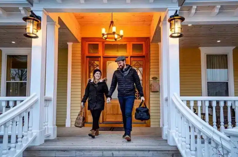 Two people walk out of a stately inn holding their overnight bags during winter.
