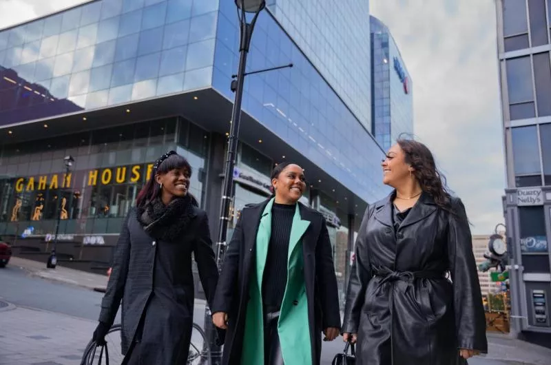 Three people walk downtown in front of a large glass building. They are wearing nice winter coats.