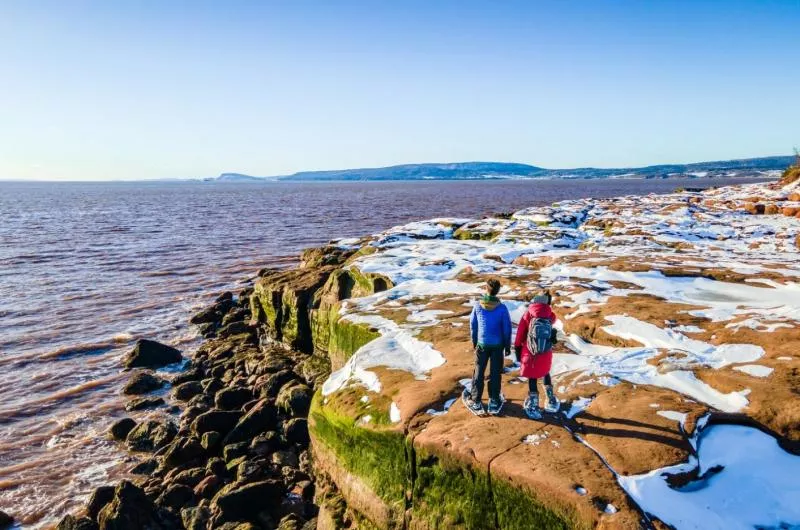 Two people, one in a blue coat and one in a red coat, stand on the edge of a rocky ocean cliff during winter.