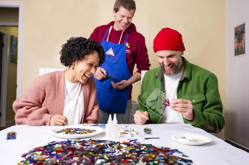 Two people are sitting at a table making art. There is a person behind them in an apron, likely the instructor, assisting.