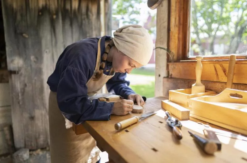A person dressed in period clothing does wood turning in a wood shop.