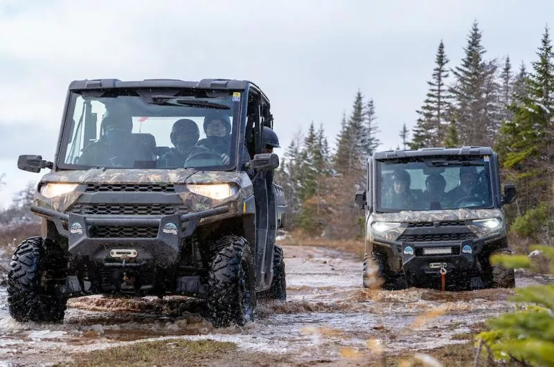 Two side-by-sides travel through a muddy trail with lots of puddles.