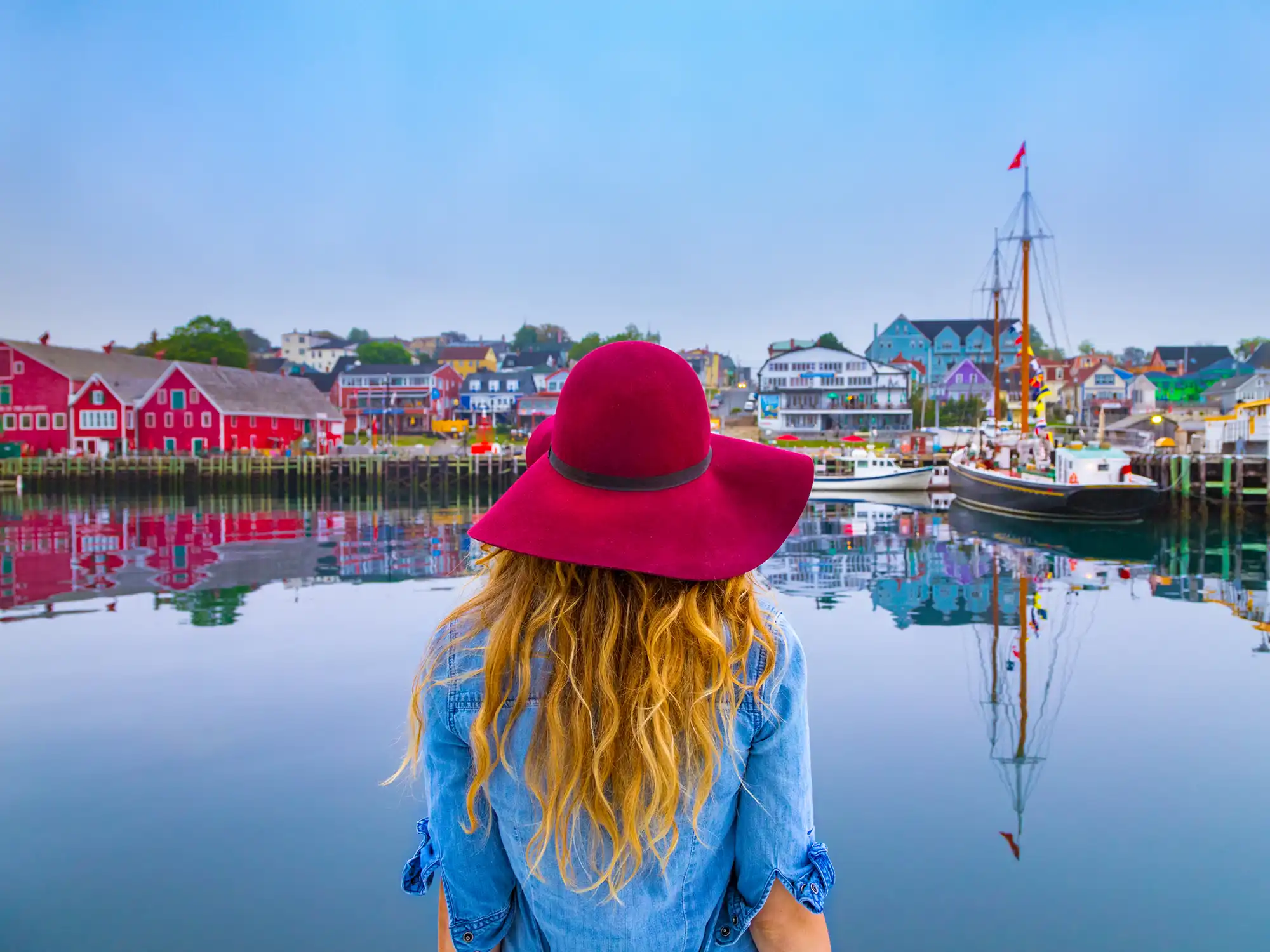 A lady in a blue shirt and red hat with blond flowy hair, stands in front of the Lunenburg waterfront which is full of colourful buildings and boats in the harbour